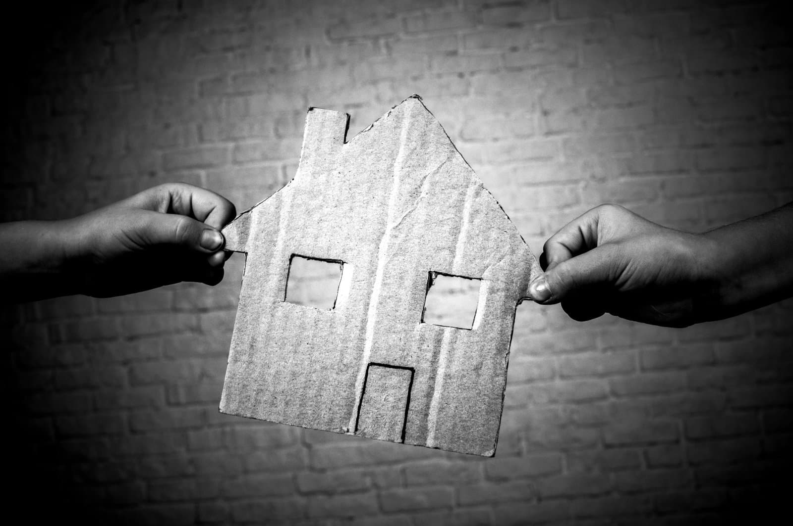 Two hands holding a cut‑out cardboard house between them against a brick wall background