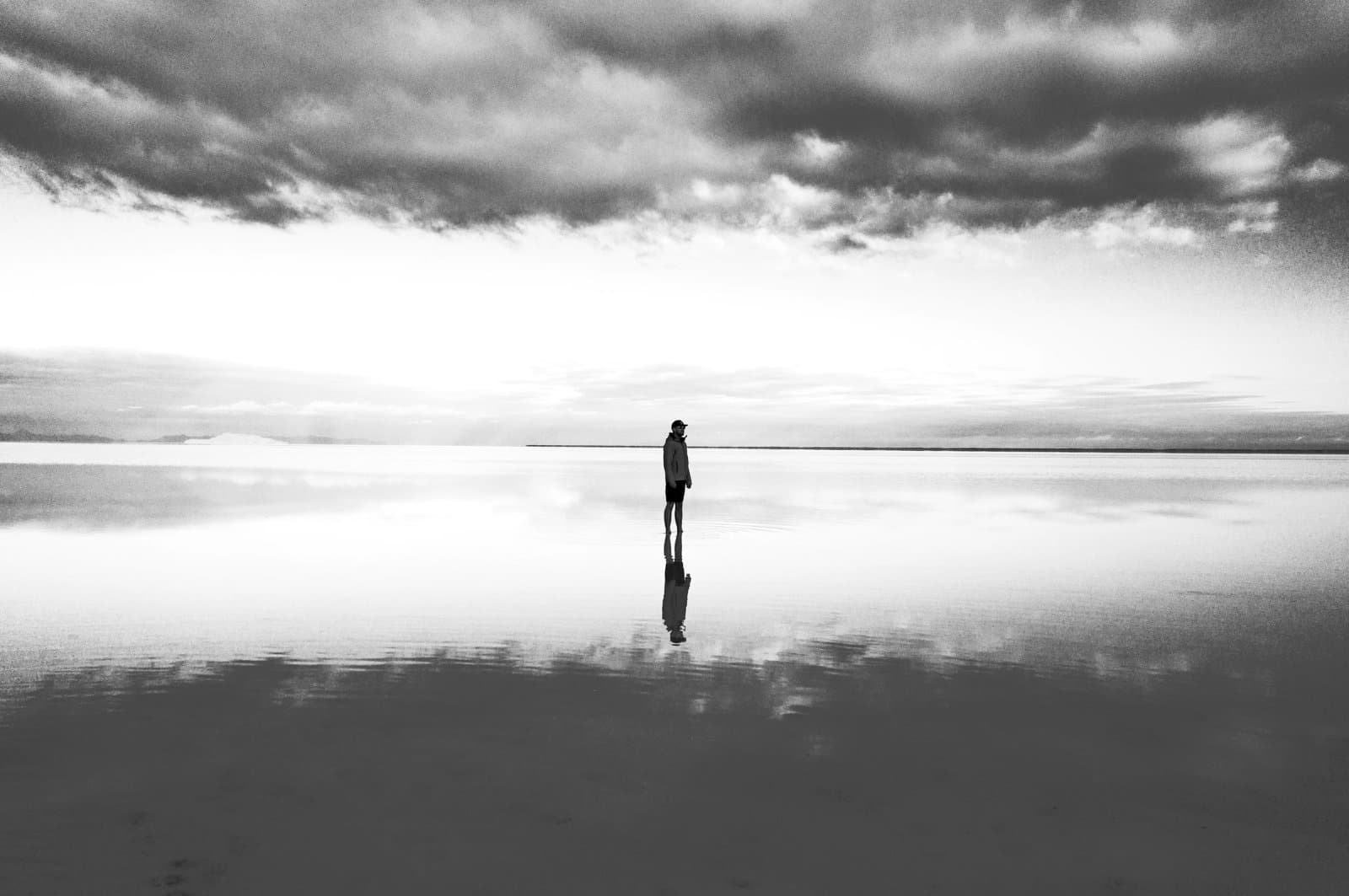 Silhouetted person standing on a reflective, shallow water surface beneath dramatic clouds.