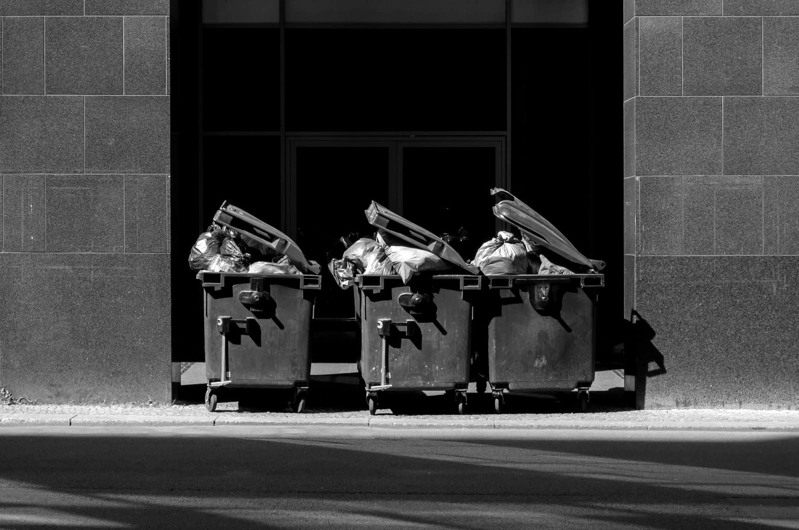 Three large black trash bins are lined up in a shaded urban alleyway, filled with blue plastic garbage bags