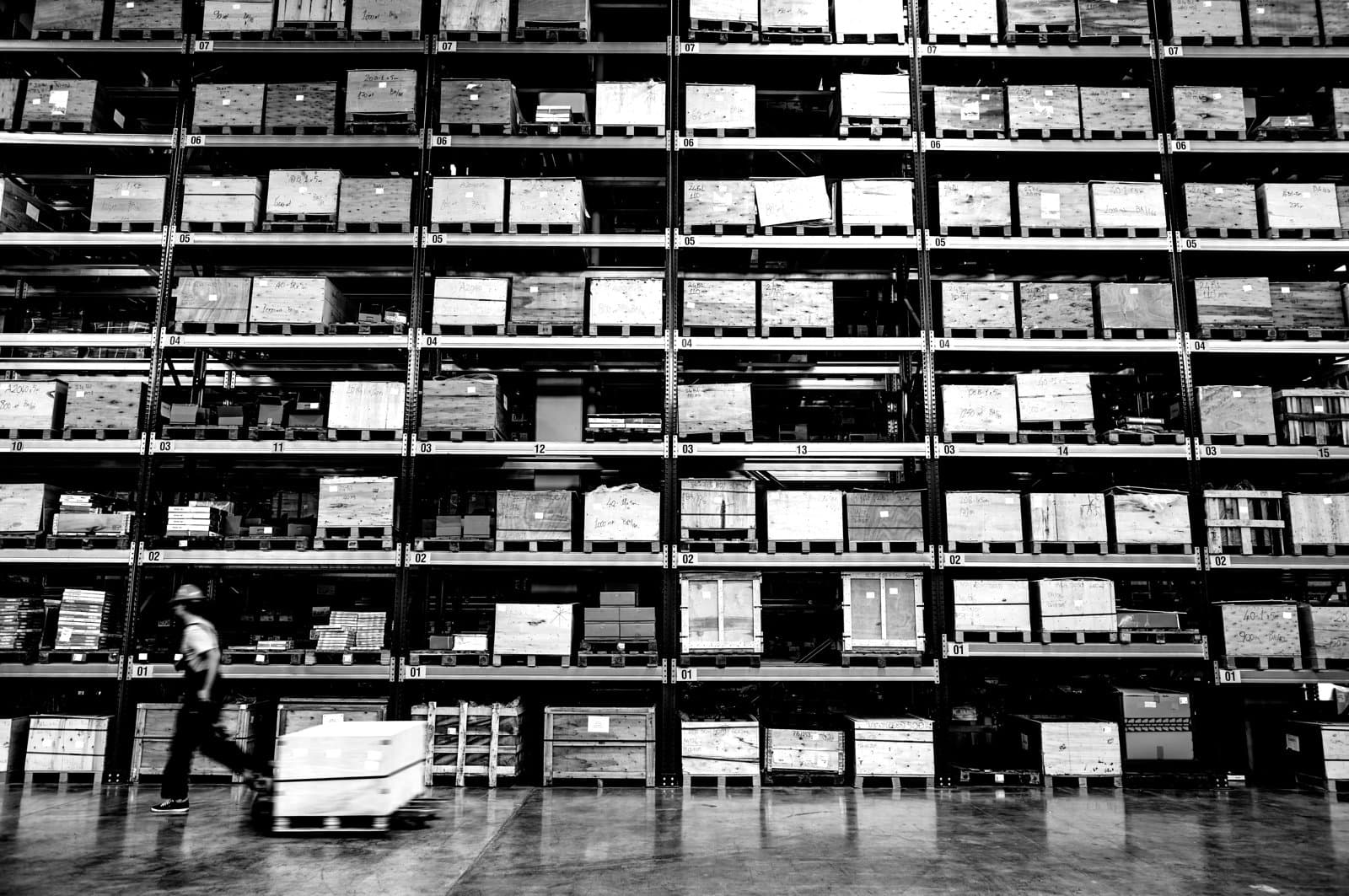 Warehouse storage racks filled with pallets and wooden crates, with a worker moving goods across the floor.